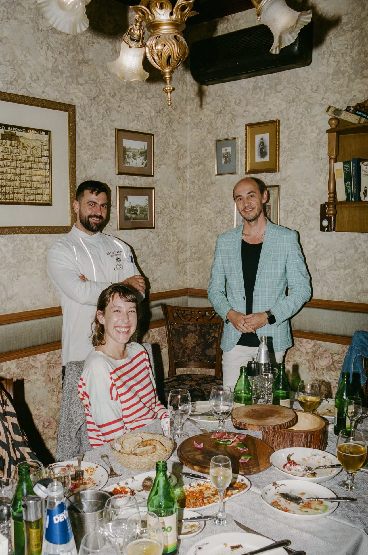 A woman (Ella) smiles at a table covered with food. Two men (the chef and the owner of Staria Chinar) stand and smile behind her