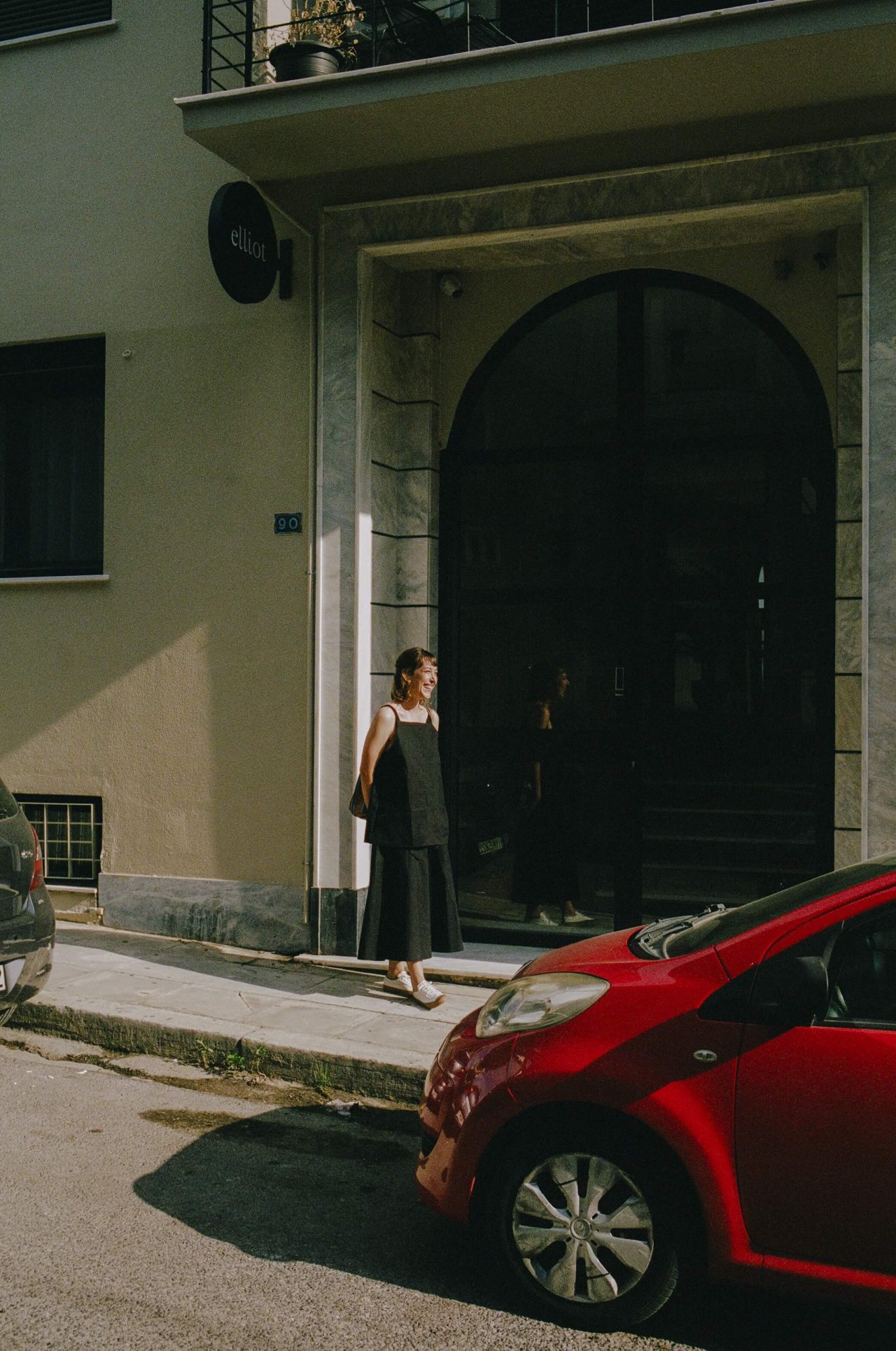 A woman in a black dress stands outside the door of a hotel, smiling into the sun