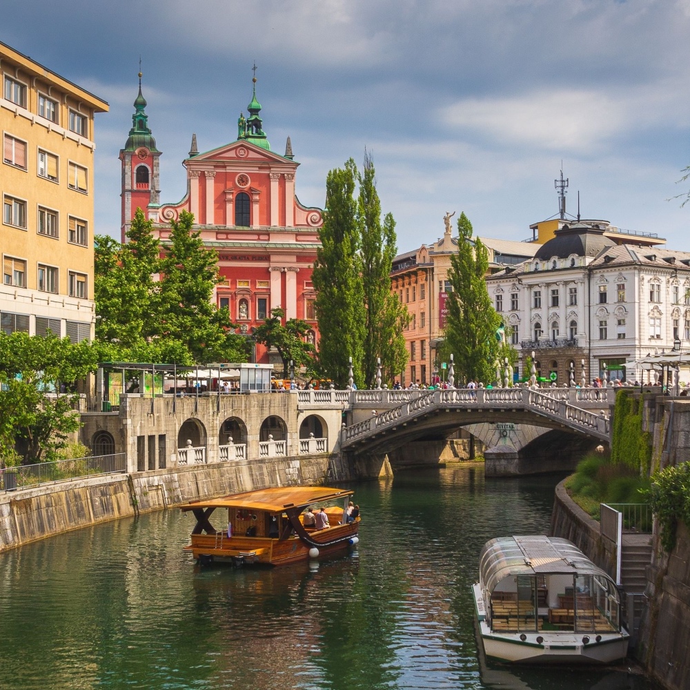 Boats glide along Ljubljana's river, with a bridge and pink church in the background